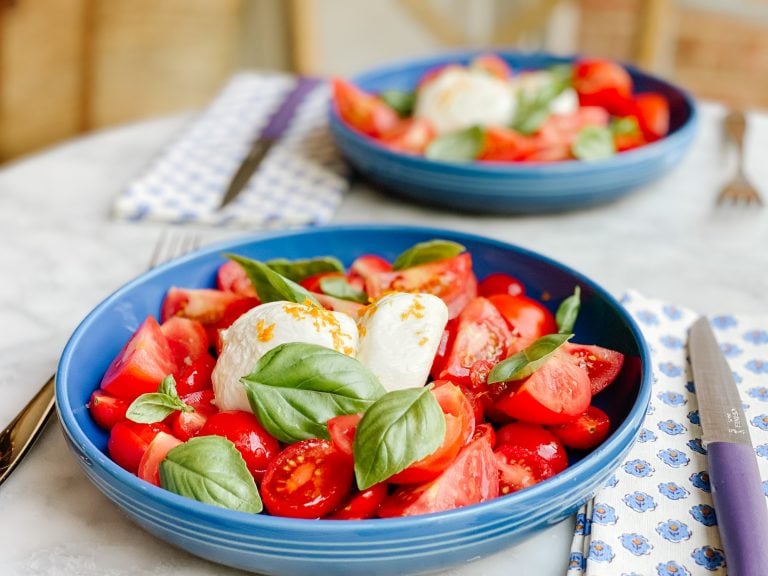 tomato basil salad in a blue bowl on a marble table with a napkin and a knife