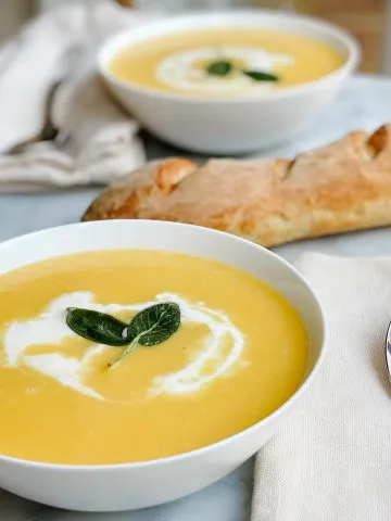 two bowls butternut squash soup with cream and sage on a table with a fresh baked baguette