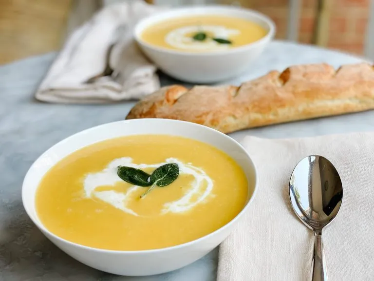 two bowls butternut squash soup with cream and sage on a table with a fresh baked baguette