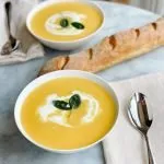 two bowls butternut squash soup with cream and sage on a table with a fresh baked baguette