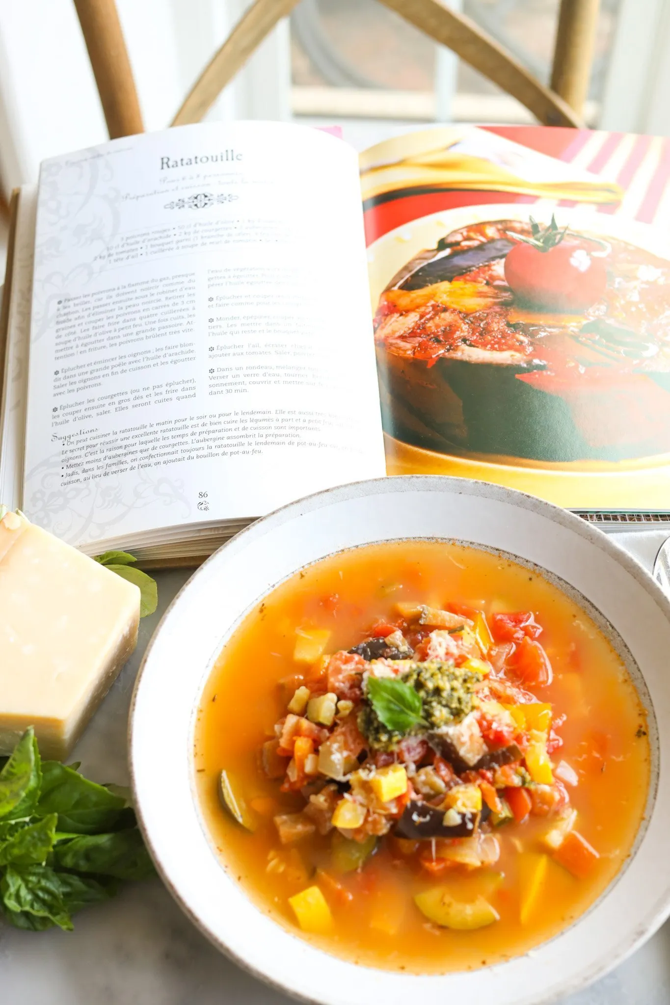 Ratatouille Soup in a bowl next to parmesan and basil with a cookbook open to a ratatouille recipe