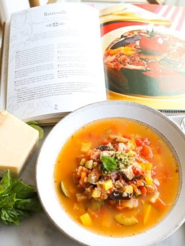 Ratatouille Soup in a bowl next to parmesan and basil with a cookbook open to a ratatouille recipe