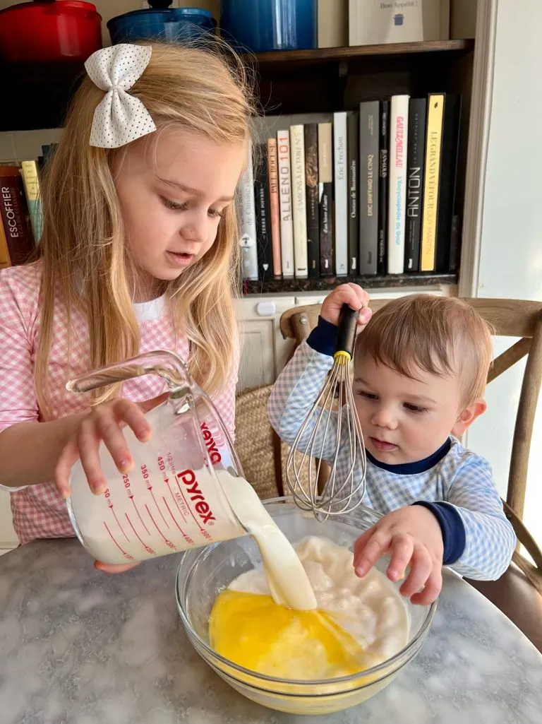 a toddler and a 19 month old cooking together - the girl pours milk into a bowl for crepes