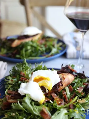 two plates of salade lyonnaise , poached egg, salad and baguette with two glasses of red wine