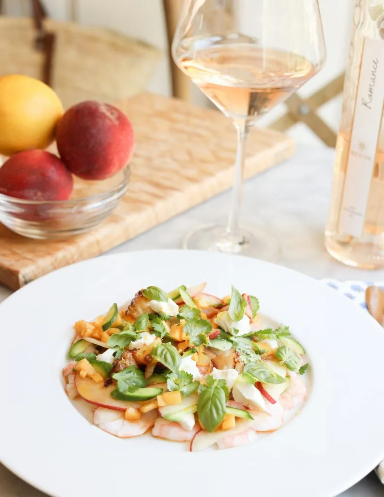 La Salade Tropezienne with shrimp, peaches, cucumbers, goat cheese, basil and cilantro and lemon thyme on a plate with a glass of Rosé wine and peaches