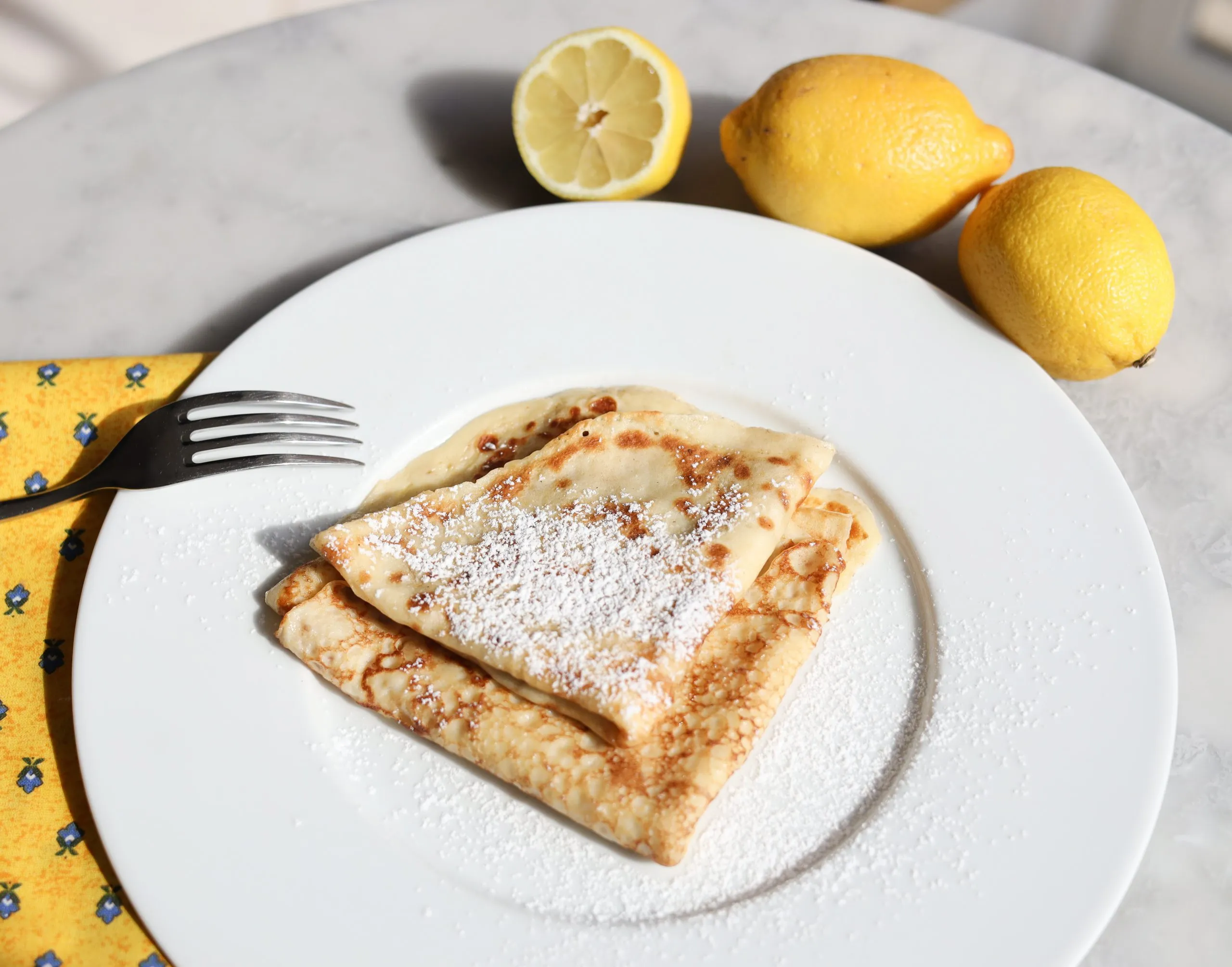 lemons surround a plate of fresh made crêpes with powdered sugar