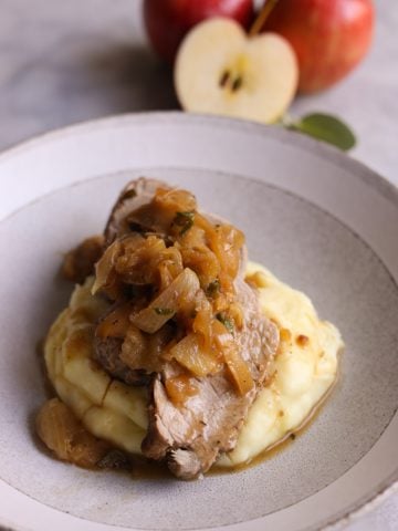 close up of a plate of mashed potatoes topped with sliced roast pork tenderloin and apples and sage