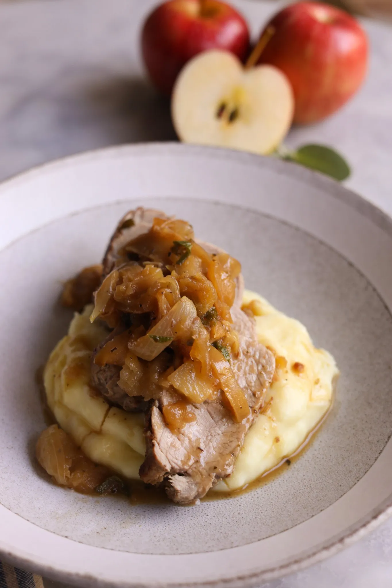 close up of a plate of mashed potatoes topped with sliced roast pork tenderloin and apples and sage