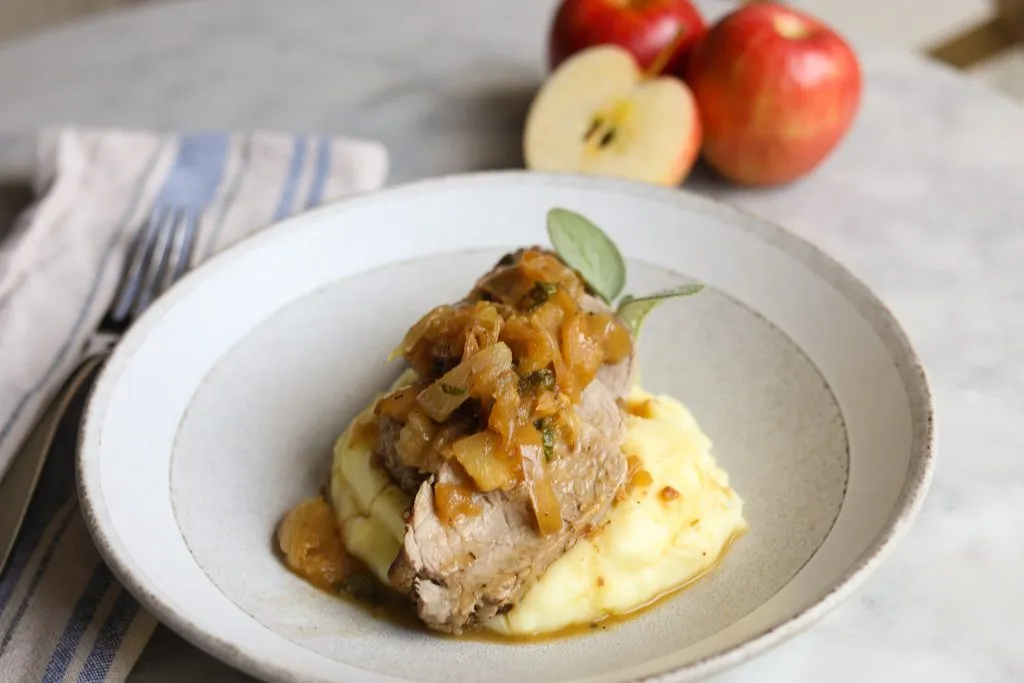 a plate of mashed potatoes topped with sliced roast pork tenderloin with apples and sage