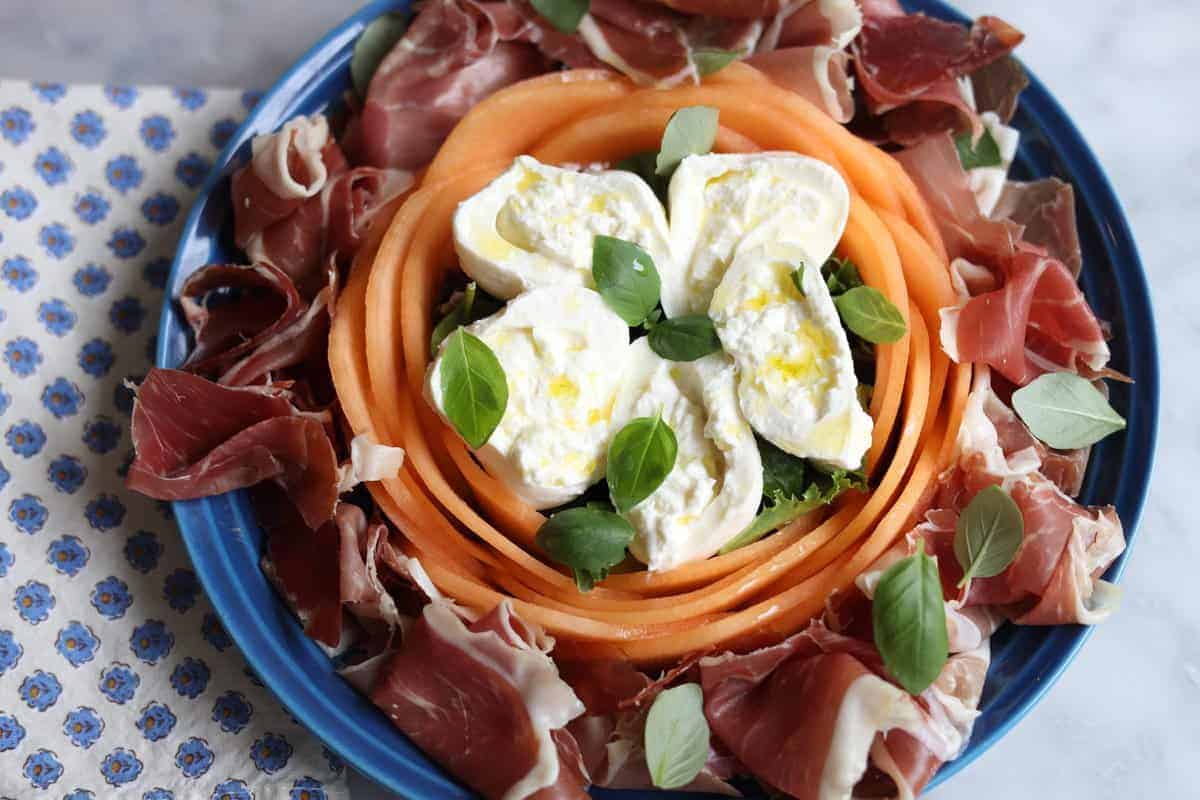 melon and burrata salad with prosciutto in a blue le creuset plate and provencal napkin