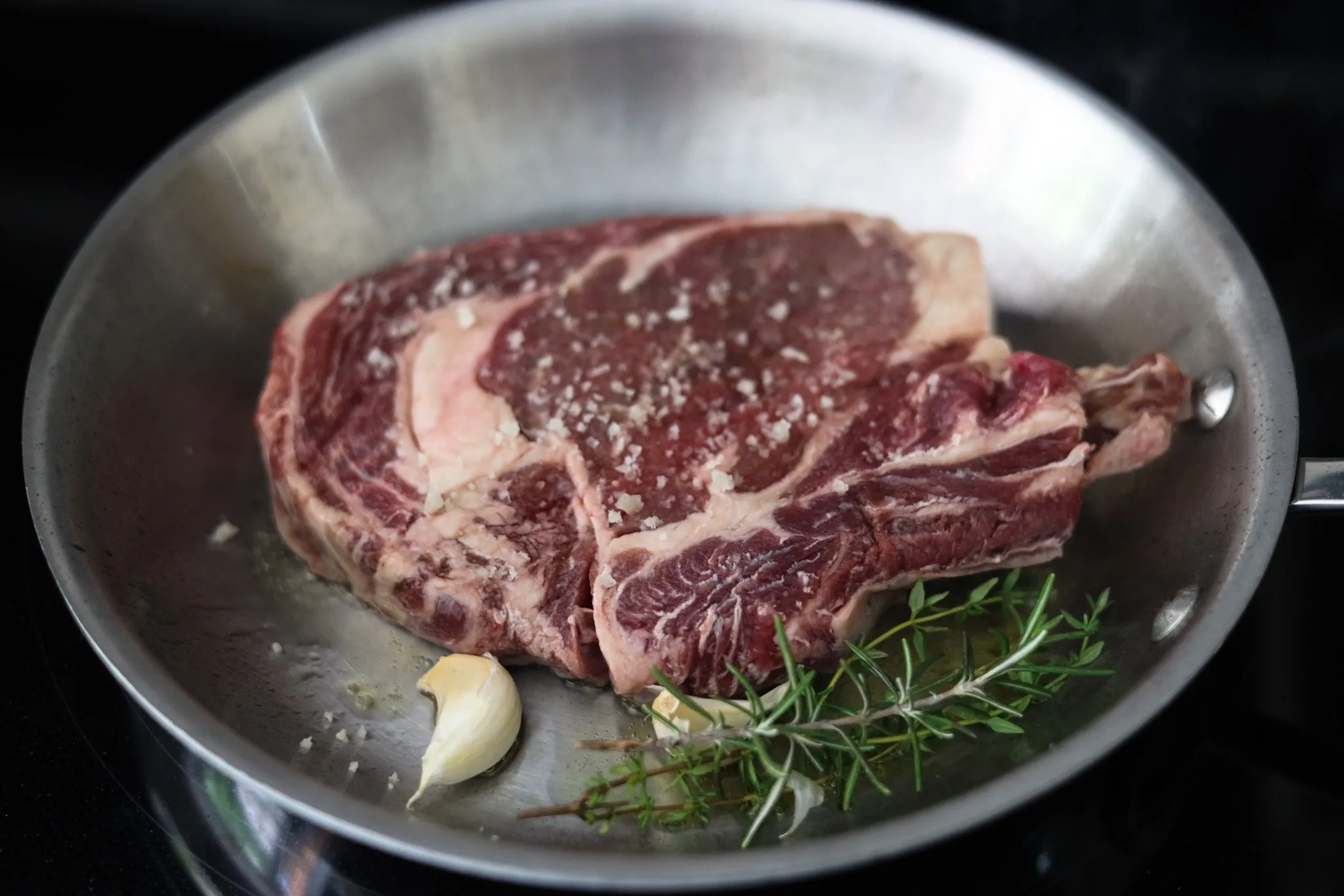a raw ribeye steak searing in the pan with fresh herbs and garlic
