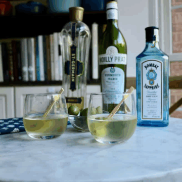 two glasses of olivette gin martini on a table with a bottle of bombay sapphire gin , noilly prat vermouth and st-germain in the background