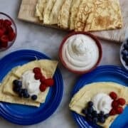 Bastille day crepes, 2 blue plates set with crepes topped with blue berries and strawberries and chantilly cream. a red bowl of chantilly cream next to crepes on a wooden cutting board