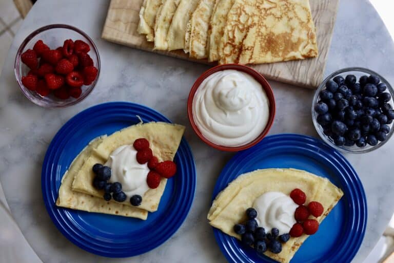 Bastille day crepes, 2 blue plates set with crepes topped with blue berries and strawberries and chantilly cream. a red bowl of chantilly cream next to crepes on a wooden cutting board