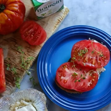 ripe summer tomatoes on a cutting board with thym, sourdough bread and boursin cheese. a tomato tartine made with the boursin cheese sits on a plate in the foreground