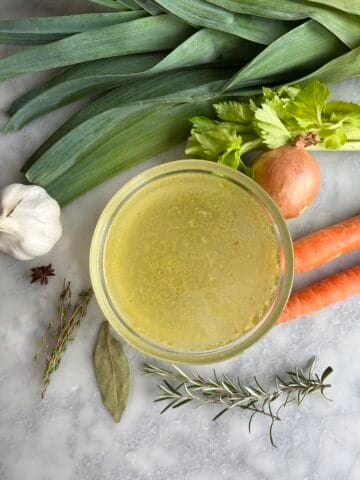 a bowl of homemade chicken stock with the ingredients besides it - leeks, onions, carrots, garlic, anise see, bay leaf, thyme, rosemary