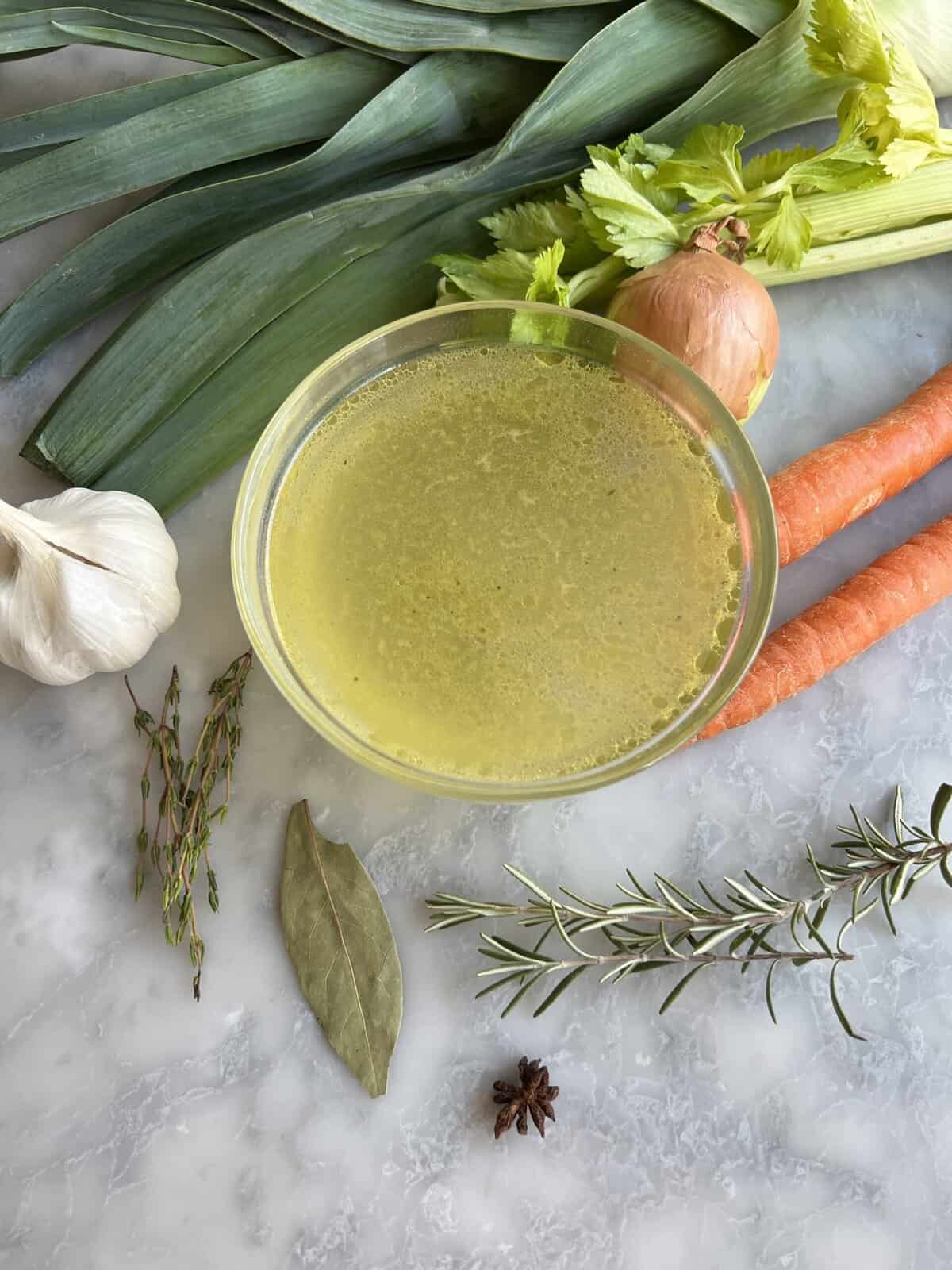 a bowl of homemade chicken stock with the ingredients besides it - leeks, onions, carrots, garlic, anise see, bay leaf, thyme, rosemary