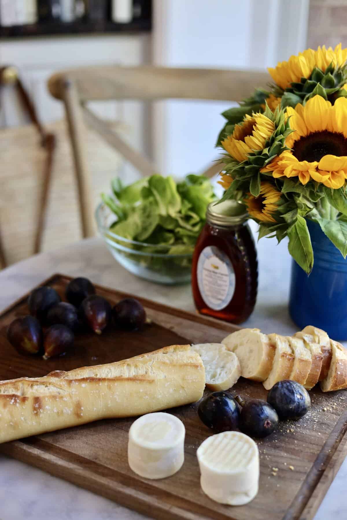 ingredients for warm goat cheese salad on a cutting board in a kitchen with sunflowers - baguette, goat cheese, figs and honey