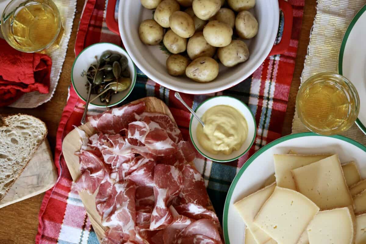 the table dressed with items for Raclette - raclette cheese, charcuterie, potatoes, capers, sourdough bread and cider