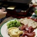 a close up of French Raclette cheese melted on potatoes with charcuterie, green salad and bread a kid pulling cheese out of a raclette grill