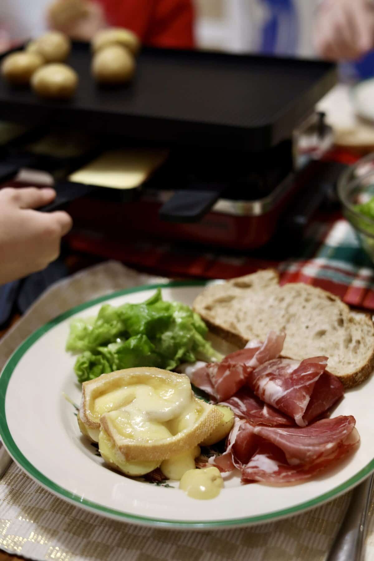 a close up of French Raclette cheese melted on potatoes with charcuterie, green salad and bread a kid pulling cheese out of a raclette grill