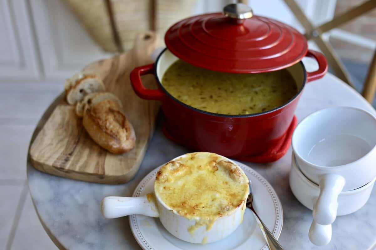 french onion soup gratinee in a bowl with melted cheese over the top next to a soup pot with onion soup inside it and a sliced baguette on the table.