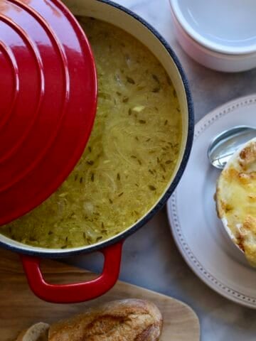french onion soup gratinee in a bowl with melted cheese over the top next to a soup pot with onion soup inside it and a sliced baguette on the table.