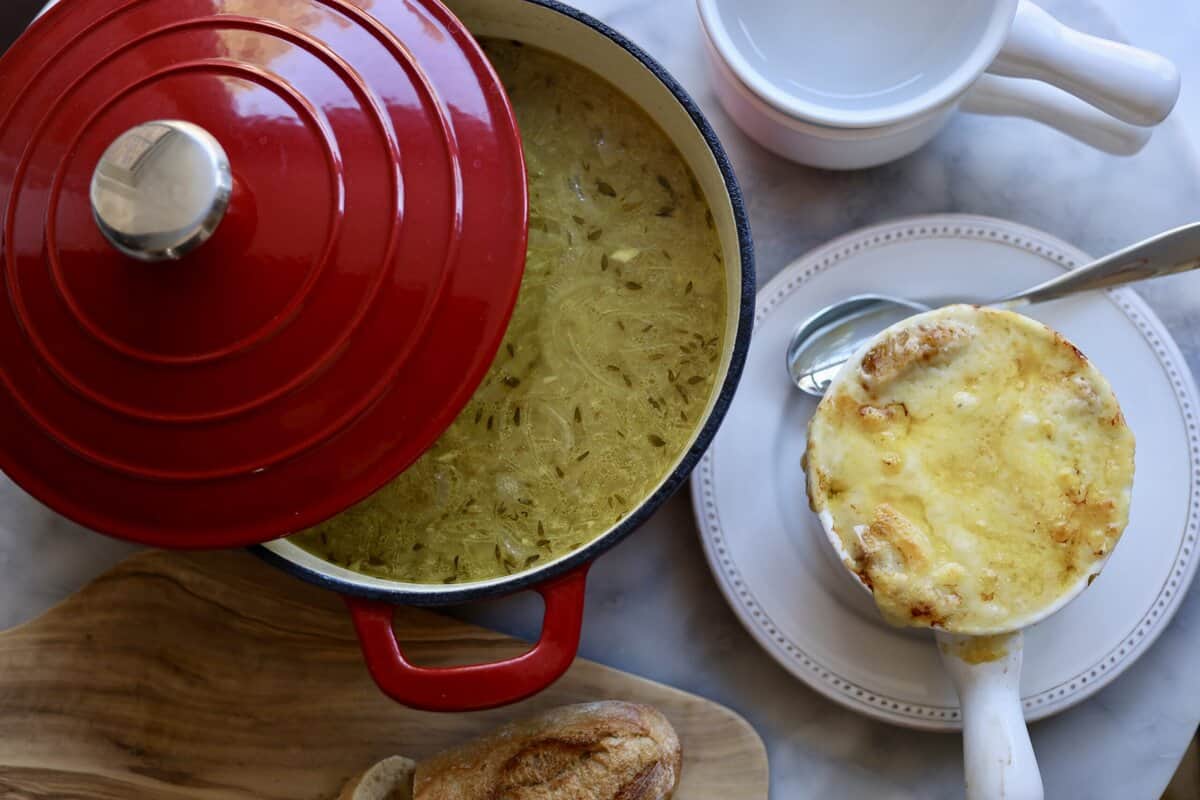 french onion soup gratinee in a bowl with melted cheese over the top next to a soup pot with onion soup inside it and a sliced baguette on the table.