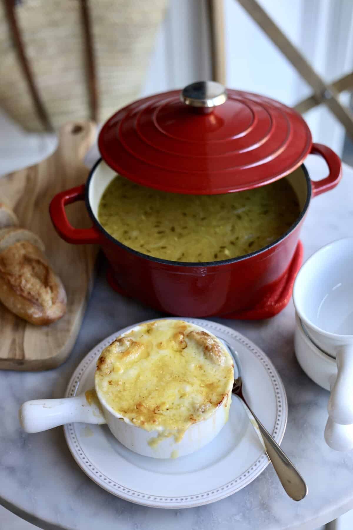 french onion soup gratinee in a bowl with melted cheese over the top next to a soup pot with onion soup inside it and a sliced baguette on the table.