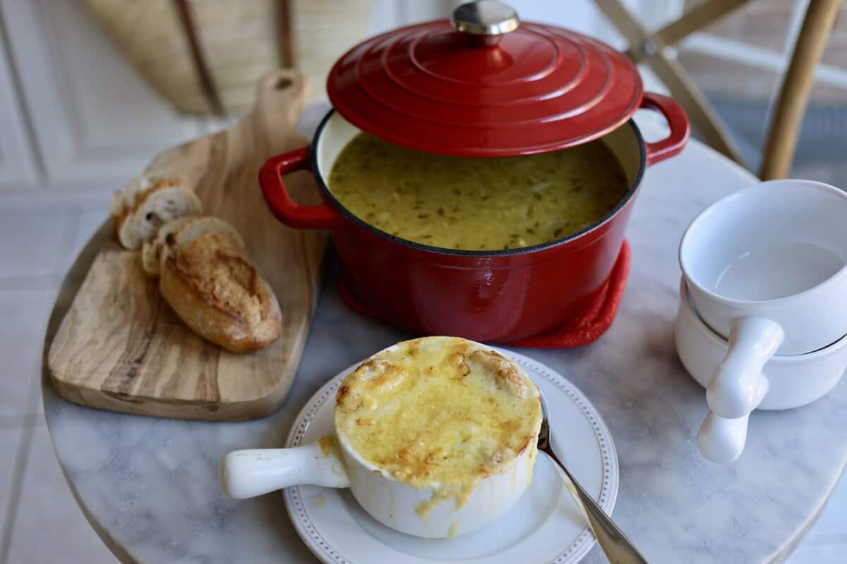 french onion soup gratinee in a bowl with melted cheese over the top next to a soup pot with onion soup inside it and a sliced baguette on the table.