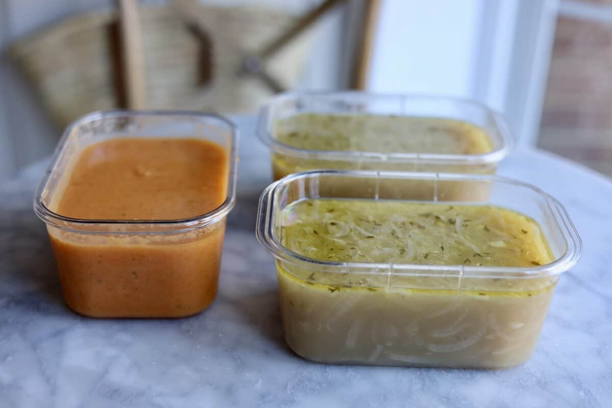 3 freezer containers with French onion soup and vegetable soup inside on a table