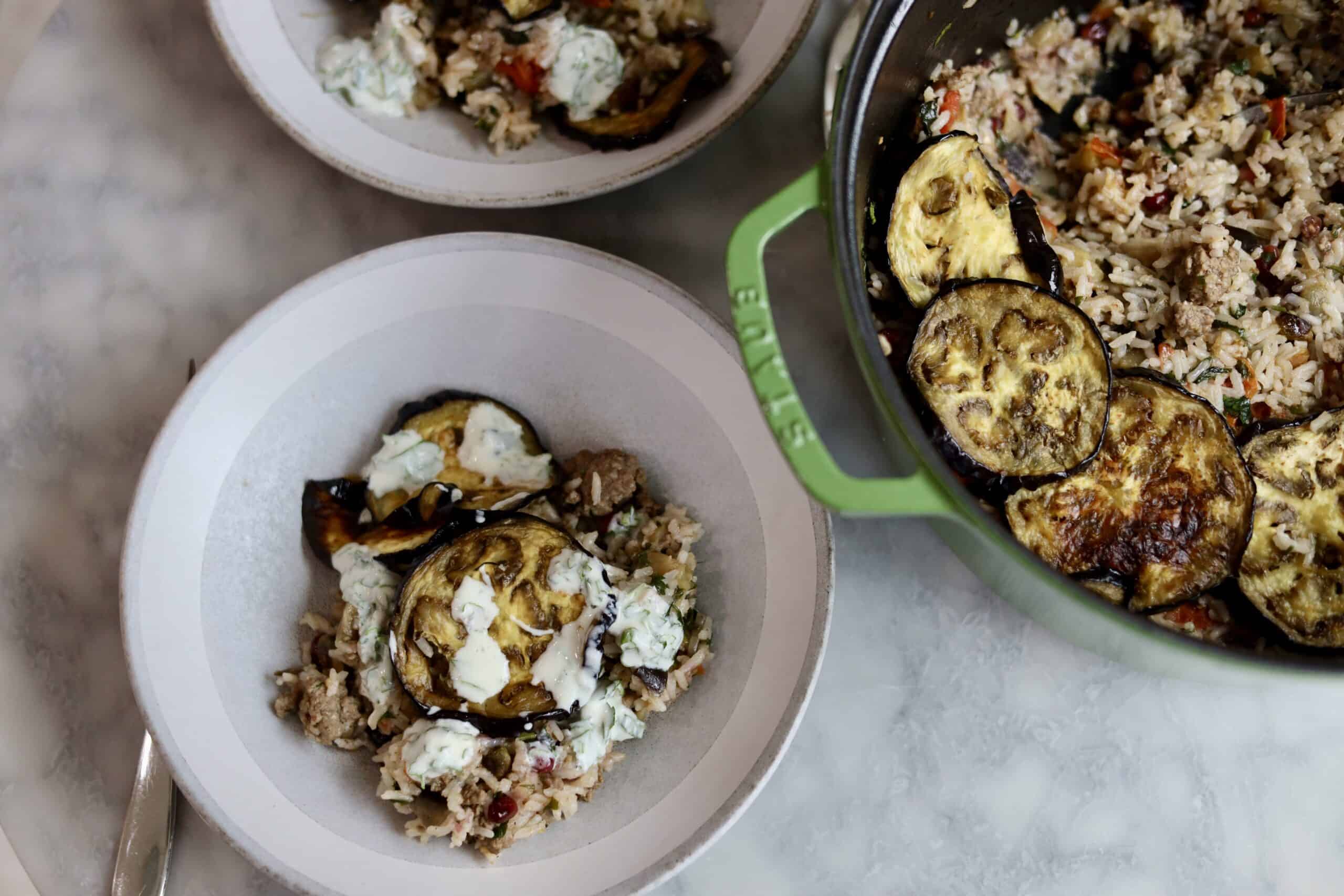 eggplant, pistachio, ground lamb, and pomegranate seed rice in a bowl with yogurt sauce next to the dutch oven it was cooked in