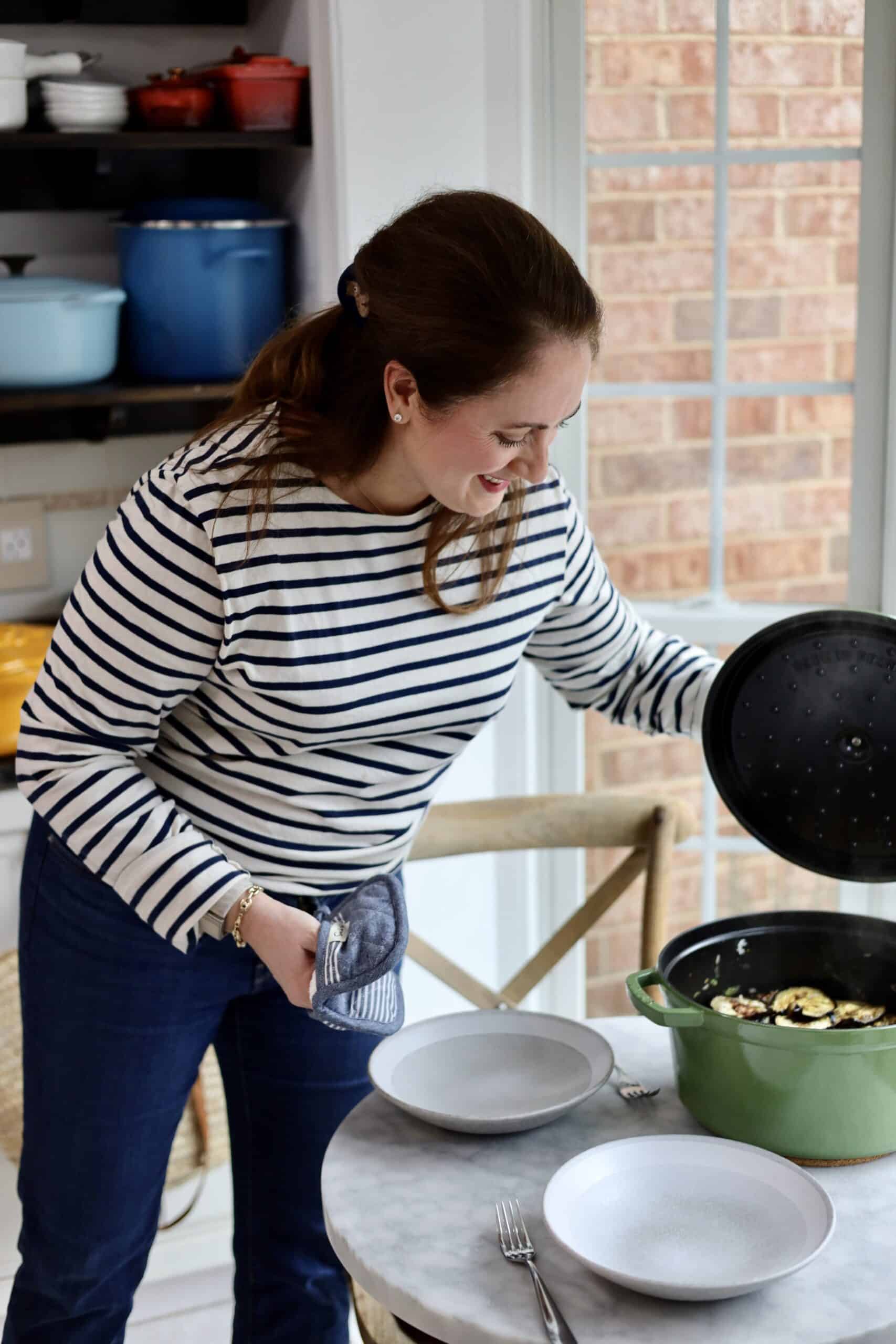 le chefs wife in her kitchen with her collection of dutch oven pots - she is wearing a striped shirt and holding a green staub pot that she set on the table next to 2 bowls