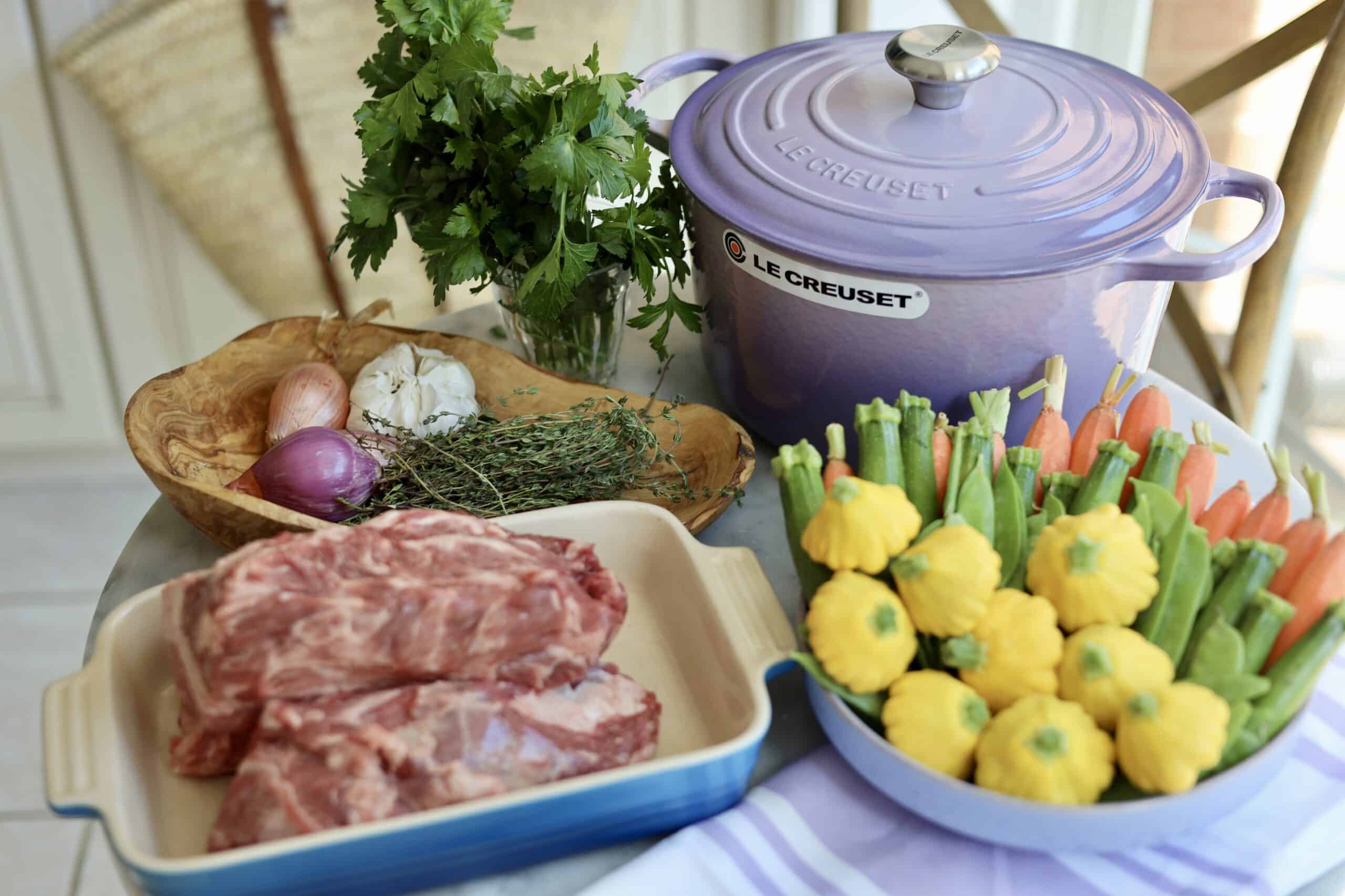 a purple hued provence Le creuset dutch oven sits on a table next to lamb meat, spring vegetables, thyme, garlic and fresh herbs