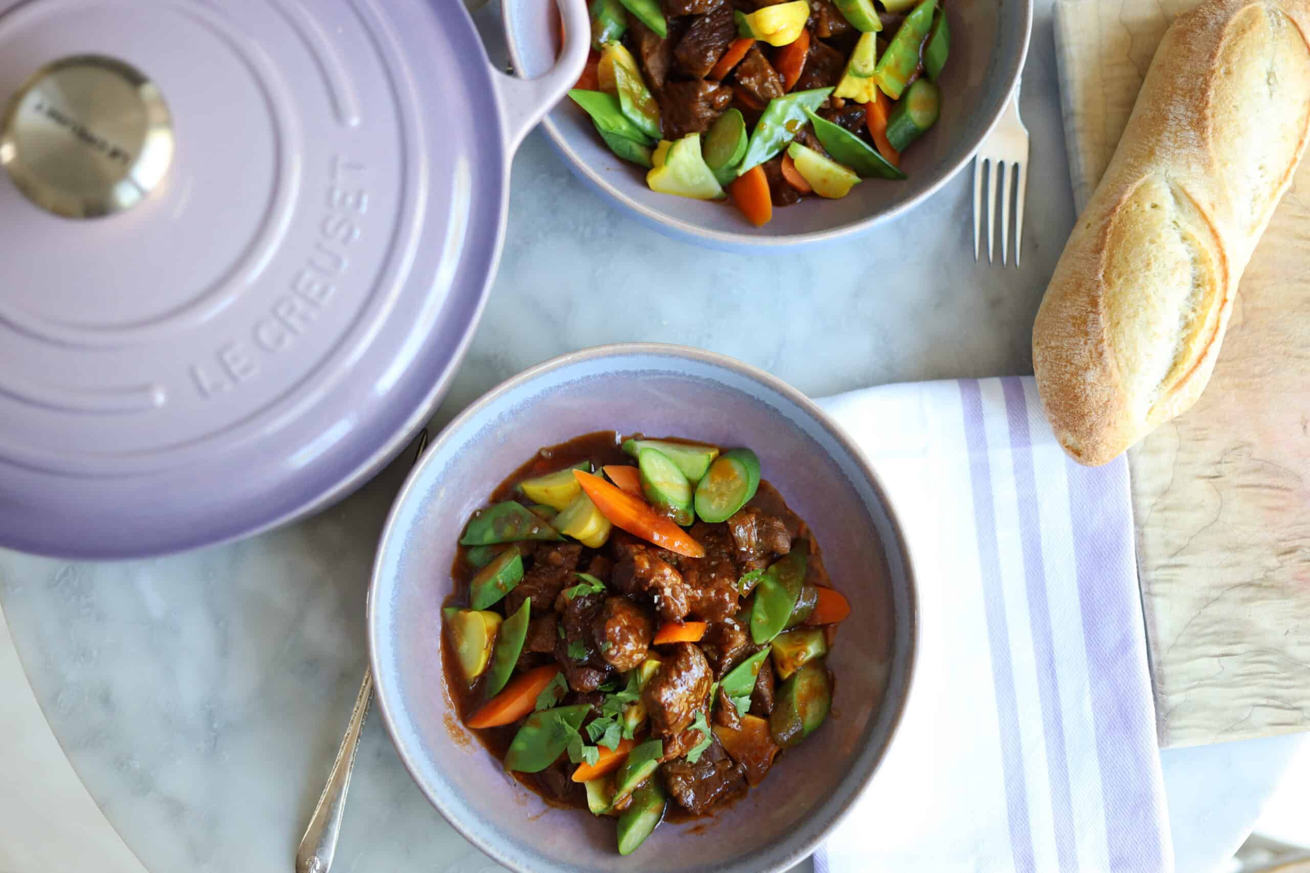 Navarin d'agneau lamb stew in a stoneware dish next to a provence purple le creuset dutch oven next to a baguette