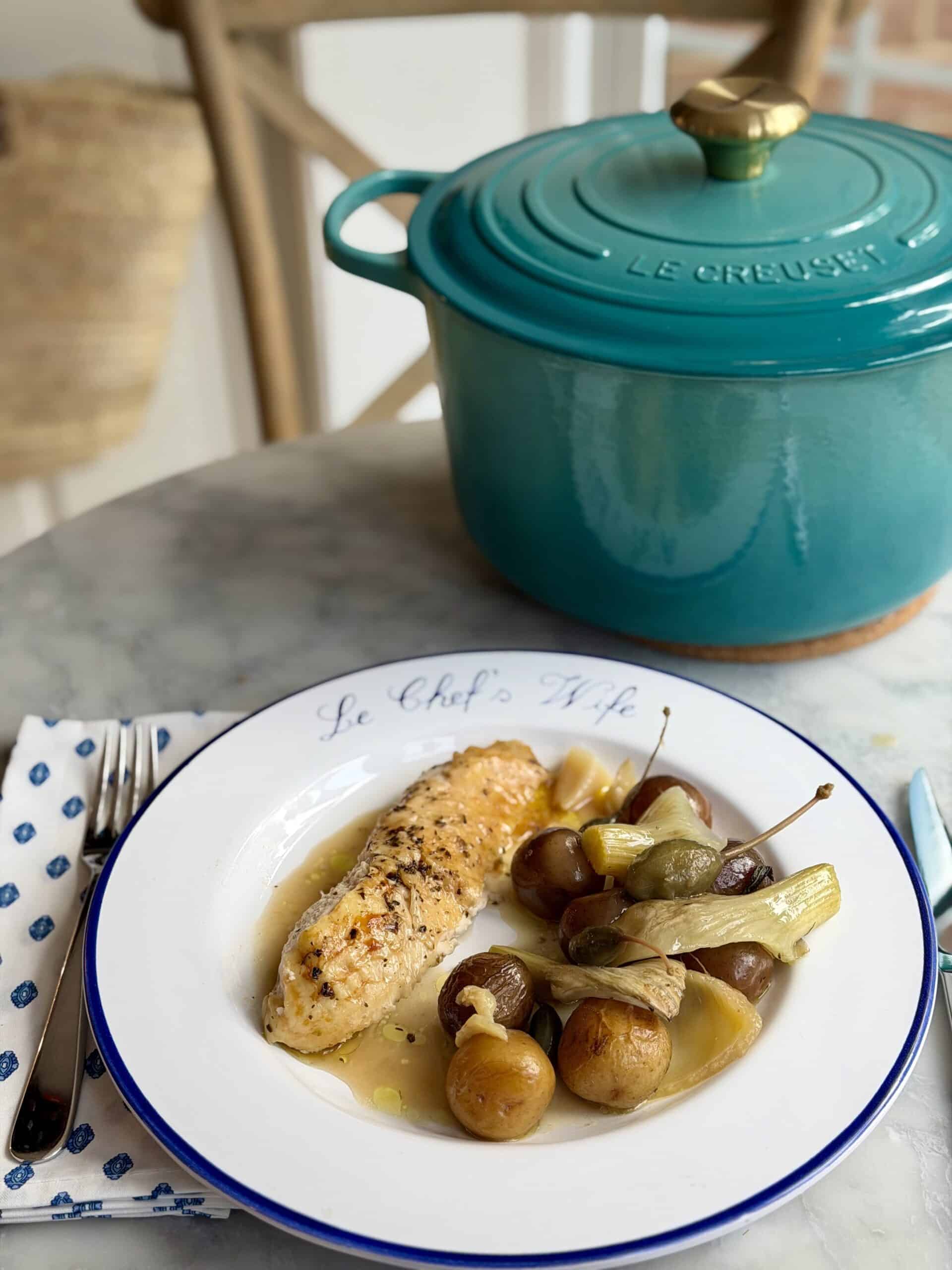 French Riviera roast chicken on a plate with a slice of chicken breast, roasted potatoes, fennel and capers behind is a Riviera colored Le Creuset Dutch Oven