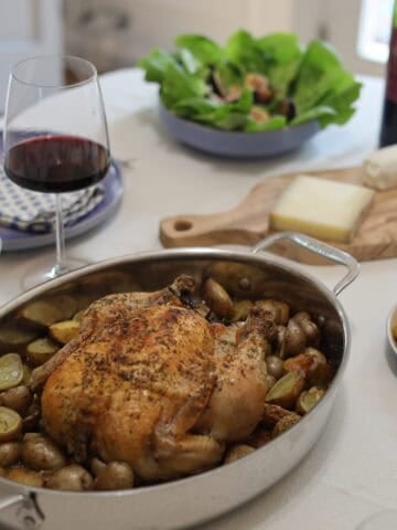a beautiful whole roast chicken with herbes de provence and herbs on a bed of potatoes fresh out of the oven on a dinner table with a glass of wine and salad and cheese platter