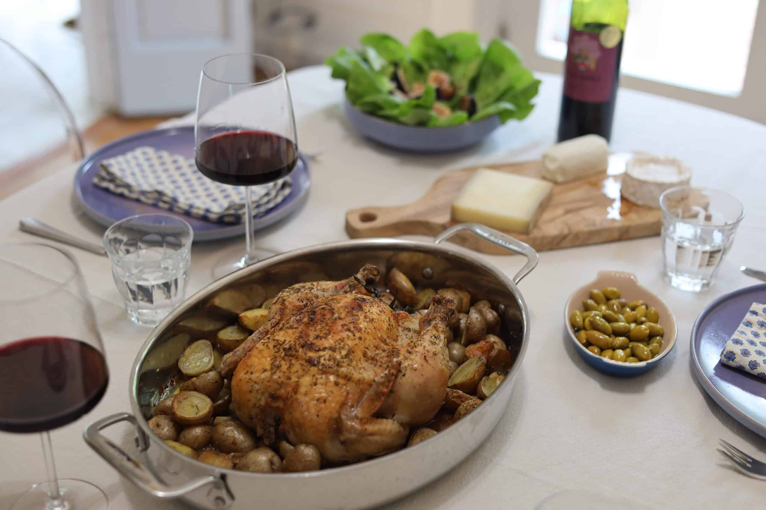 a beautiful whole roast chicken with herbes de provence and herbs on a bed of potatoes fresh out of the oven on a dinner table with a glass of wine and salad and cheese platter