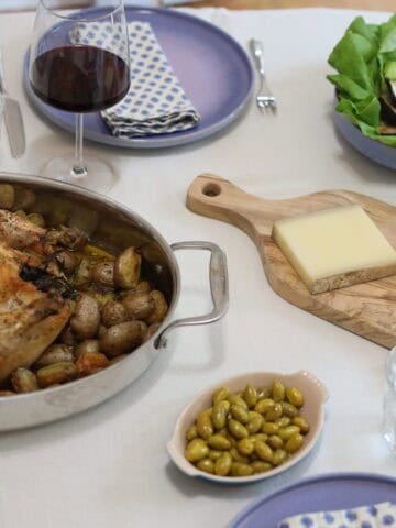 a beautiful whole roast chicken with herbes de provence and herbs on a bed of potatoes fresh out of the oven on a dinner table with a glass of wine and salad