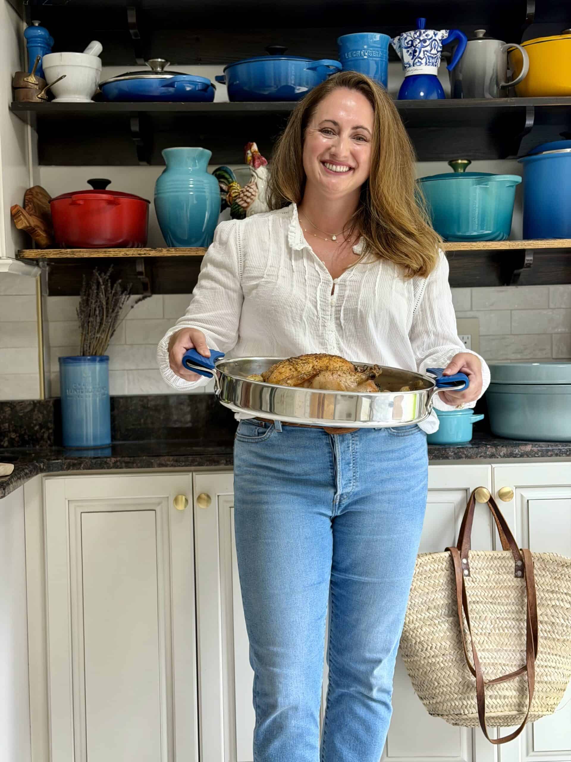 Anina Belle Giannini standing with a roast chicken in her kitchen in front of her wall of dutch ovens